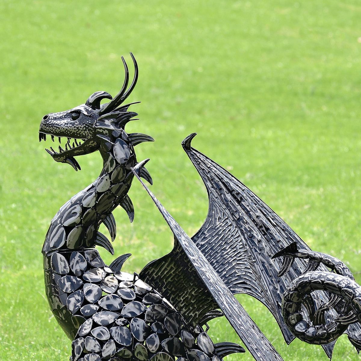 Close view of a dark metal dragon statue with a raised head, textured scales and outstretched wings against green grass.