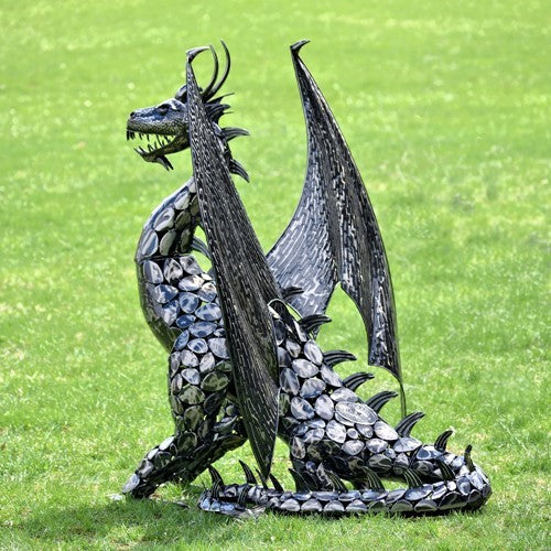 A dark metal dragon statue with raised wings sits on grass, its body covered in overlapping scales.