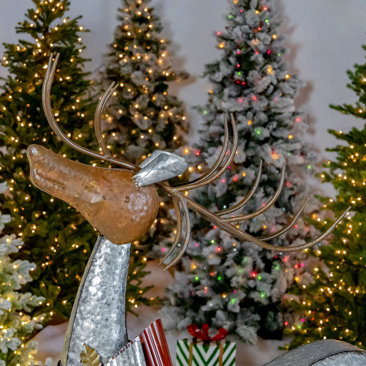 Galvanized reindeer with curved metal antlers posed before lit Christmas trees and wrapped gifts.