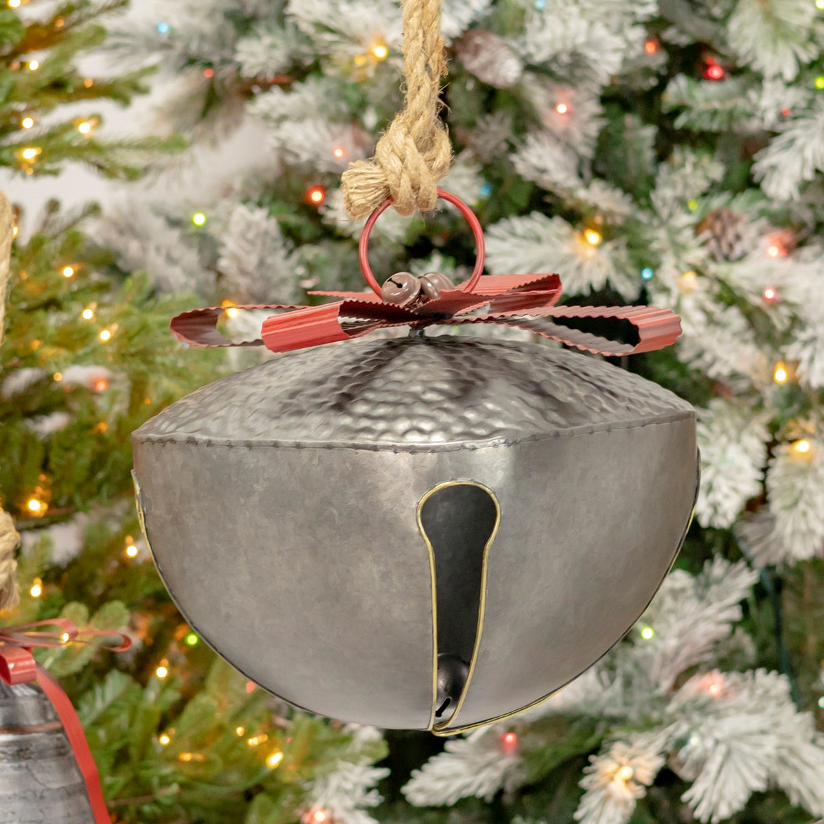 Large rounded galvanized bell with a red ribbon and rope hanger against a lit Christmas tree.