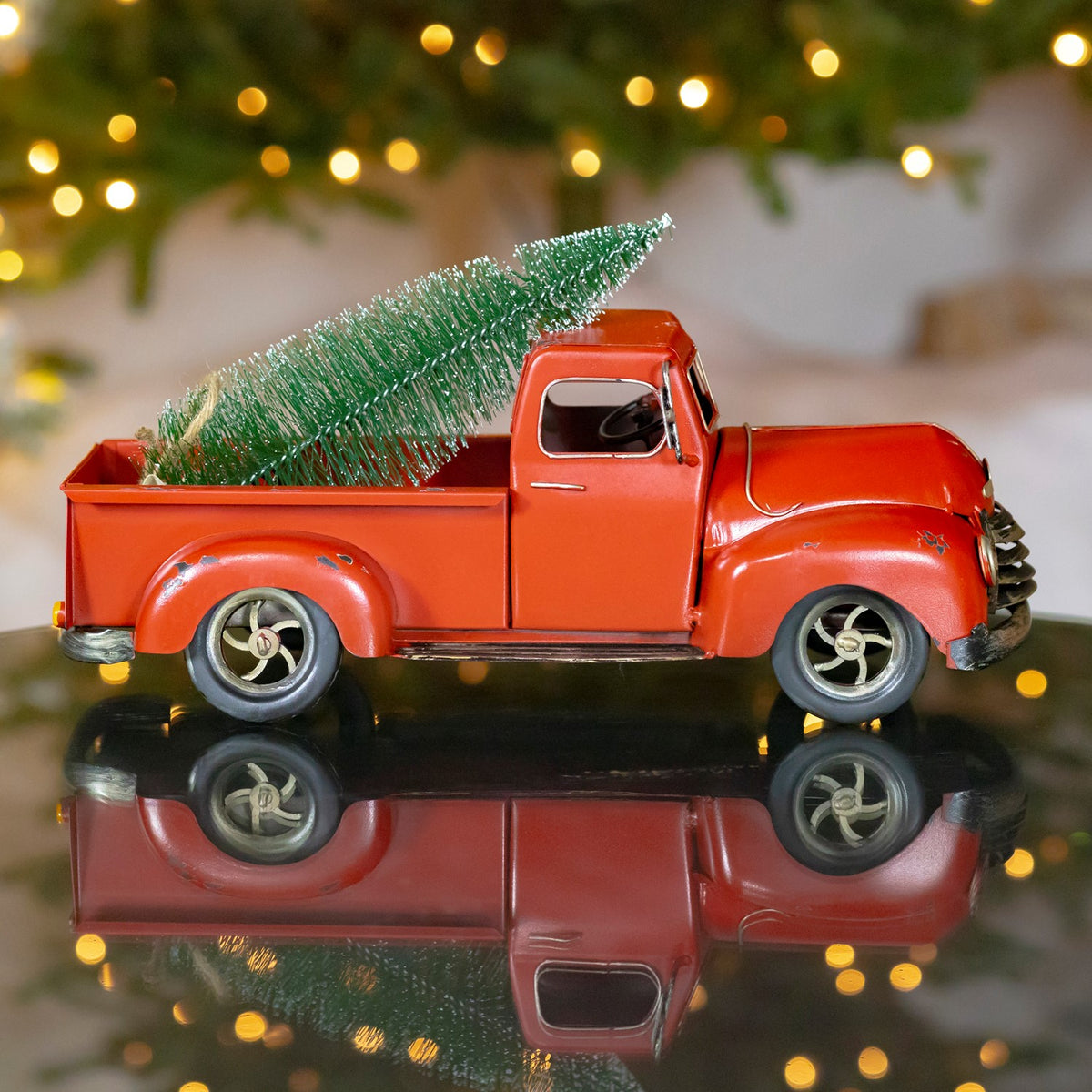 Red vintage-style pickup carrying a green tree, reflected on a glossy surface beneath festive lights.