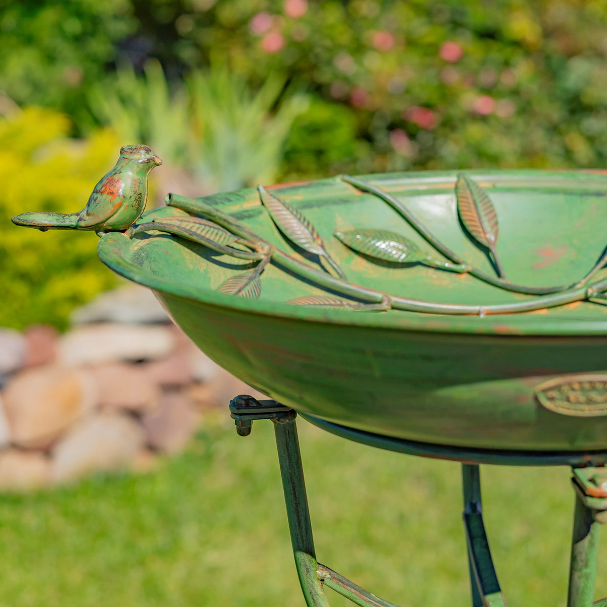 Close view of a small iron bird perched on the leaf-decorated rim of a green birdbath.