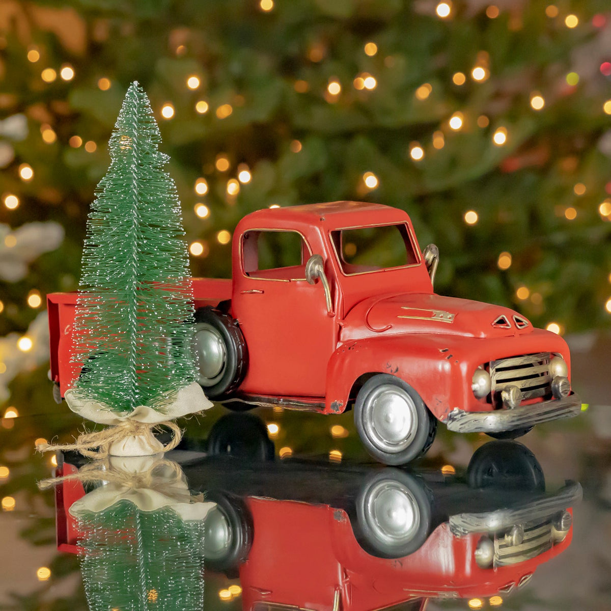 Distressed red pickup truck carrying a tall green tree, reflected on a glossy surface before a lit Christmas tree.