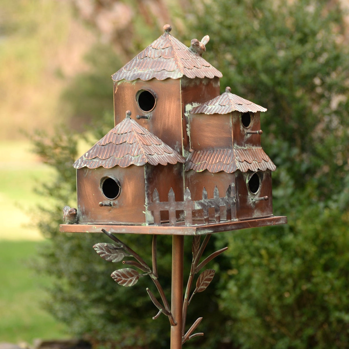 Rustic metal birdhouse stake with several roofed houses and leafy branches against a garden backdrop.