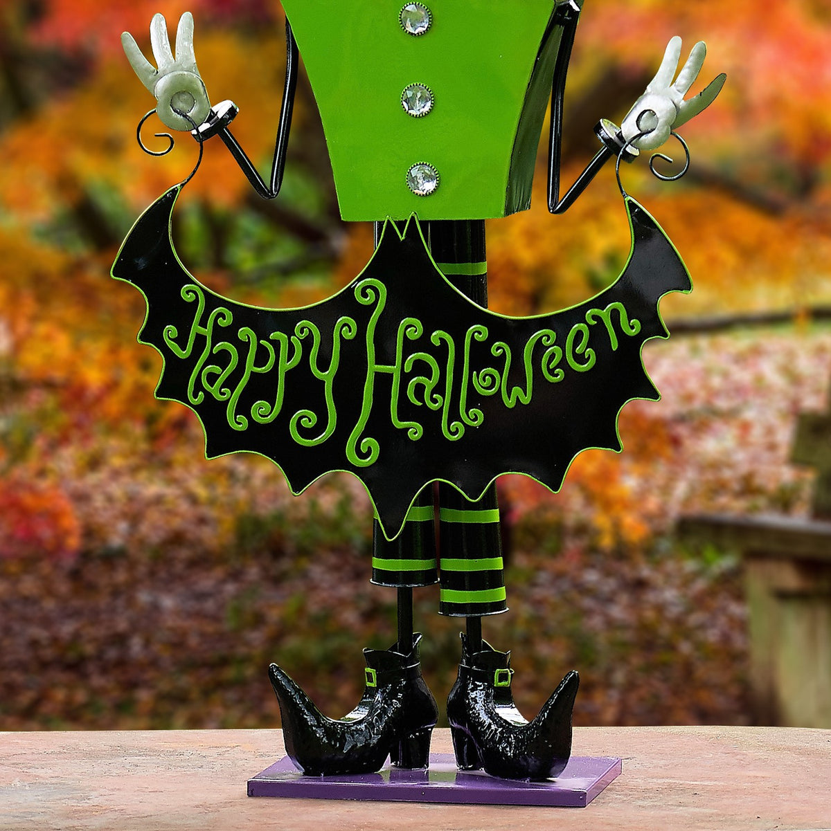 Close view of a green-costumed skeleton figurine holding a black bat-shaped Halloween sign above its boots.