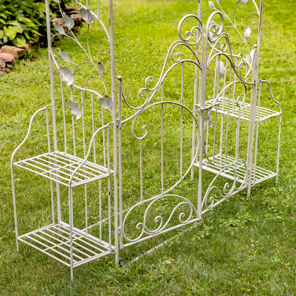 Light-colored wrought-metal garden arch with decorative gate and two-tier plant stands on grass.