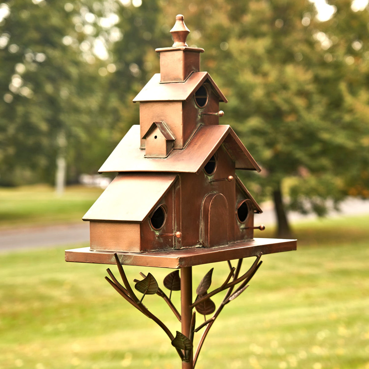Copper-toned church-shaped birdhouse on a decorative metal stake in a garden.