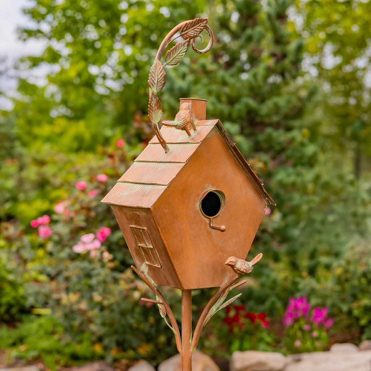 Angled view of the copper birdhouse stake with roof, bird details, and garden backdrop