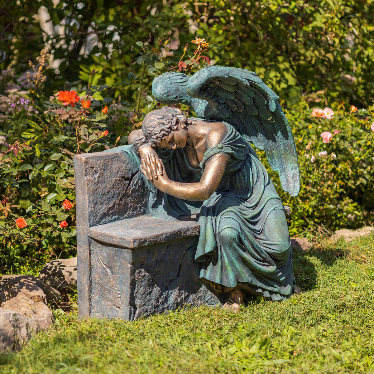 A bronze-toned angel with green patina rests her head and arms on a stone-look bench among flowers.