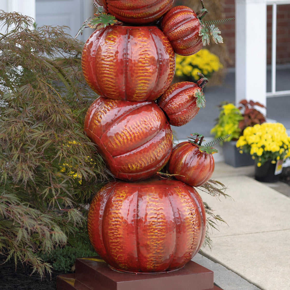 Close view of glossy orange pumpkins stacked on a square base, with foliage and yellow flowers nearby.