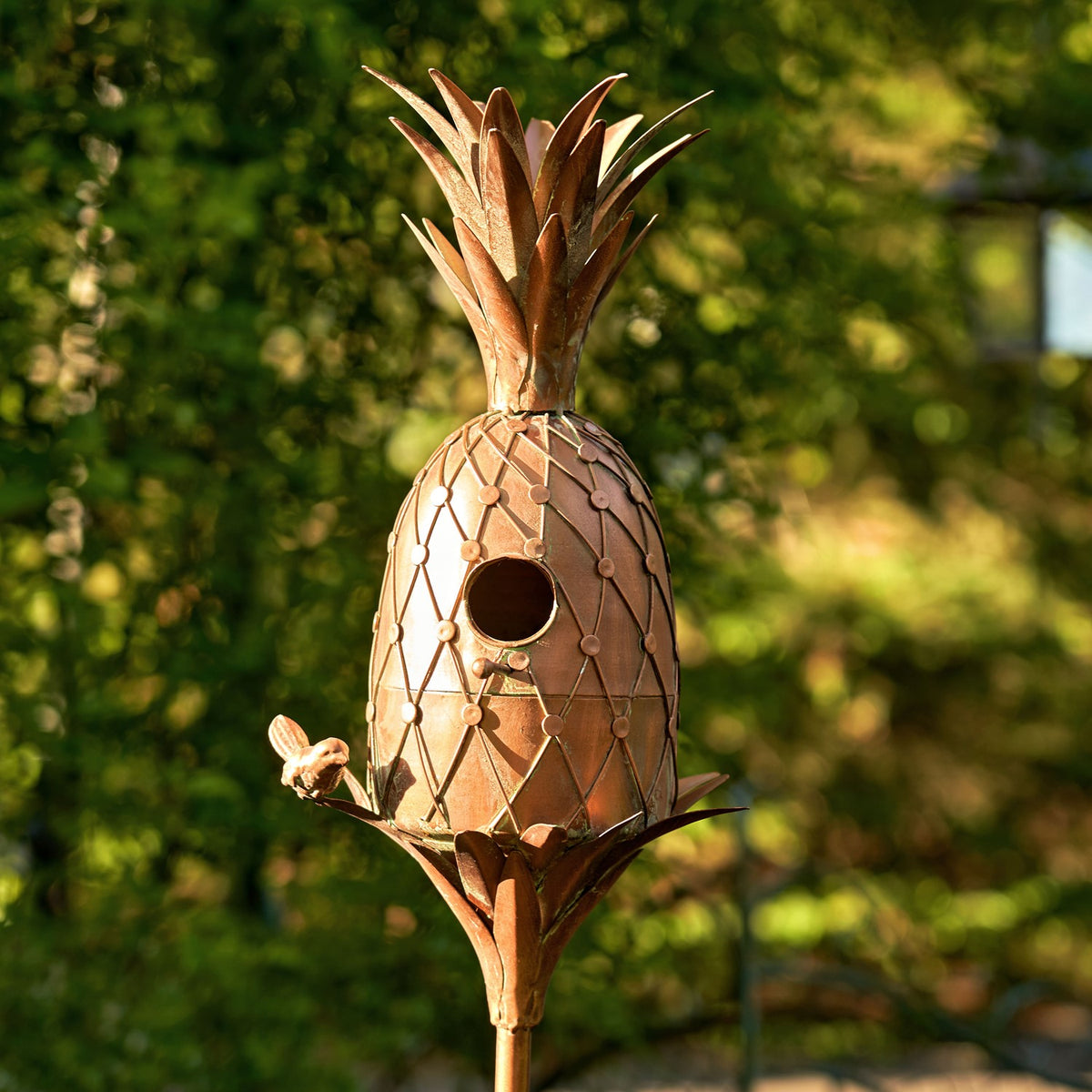Copper-tone pineapple birdhouse with entry hole, viewed against greenery