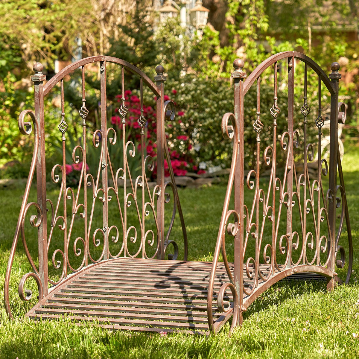 Ornate arched metal garden bridge with curled railings and slatted walkway on a lawn
