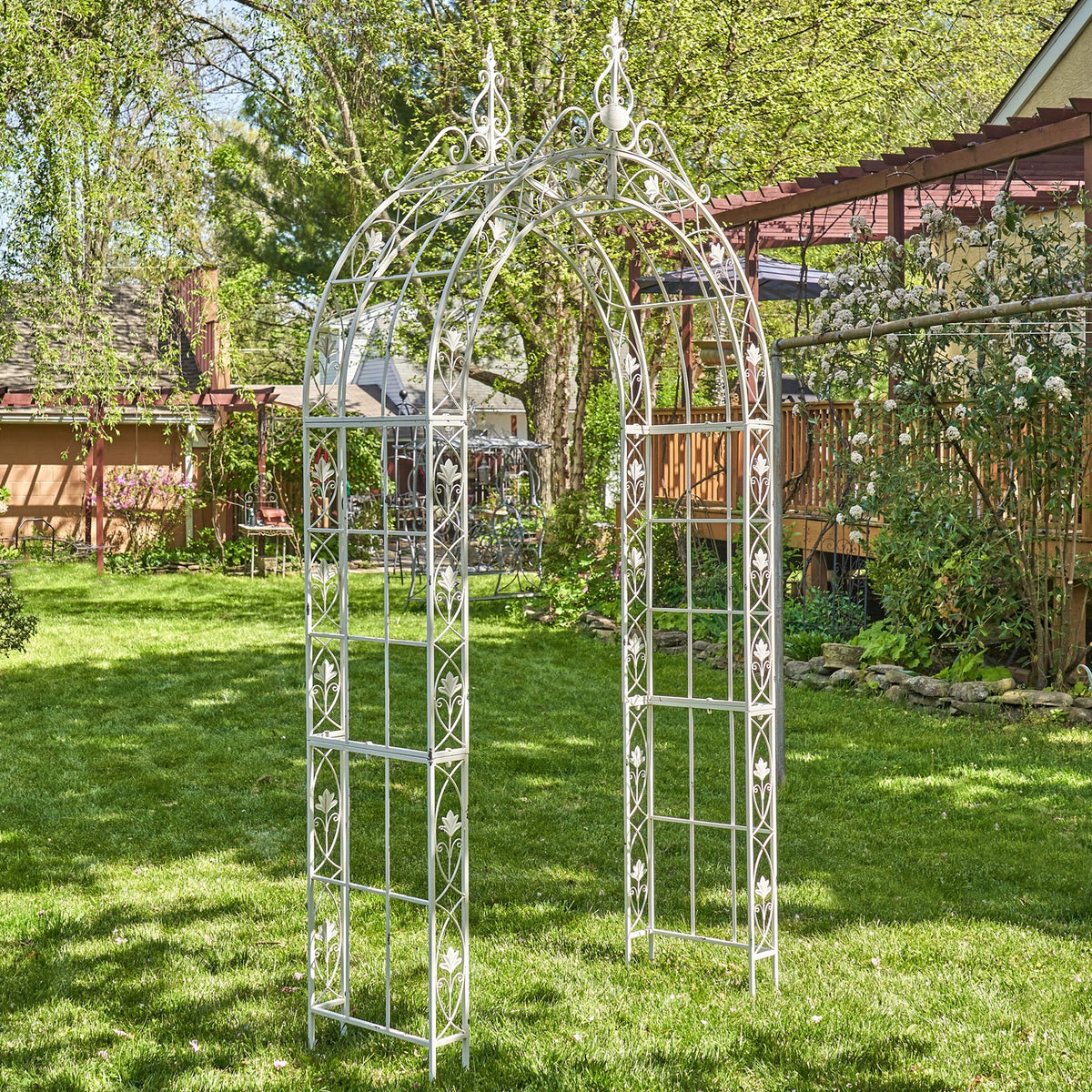 Pale metal garden arch standing on a lawn beneath leafy trees.
