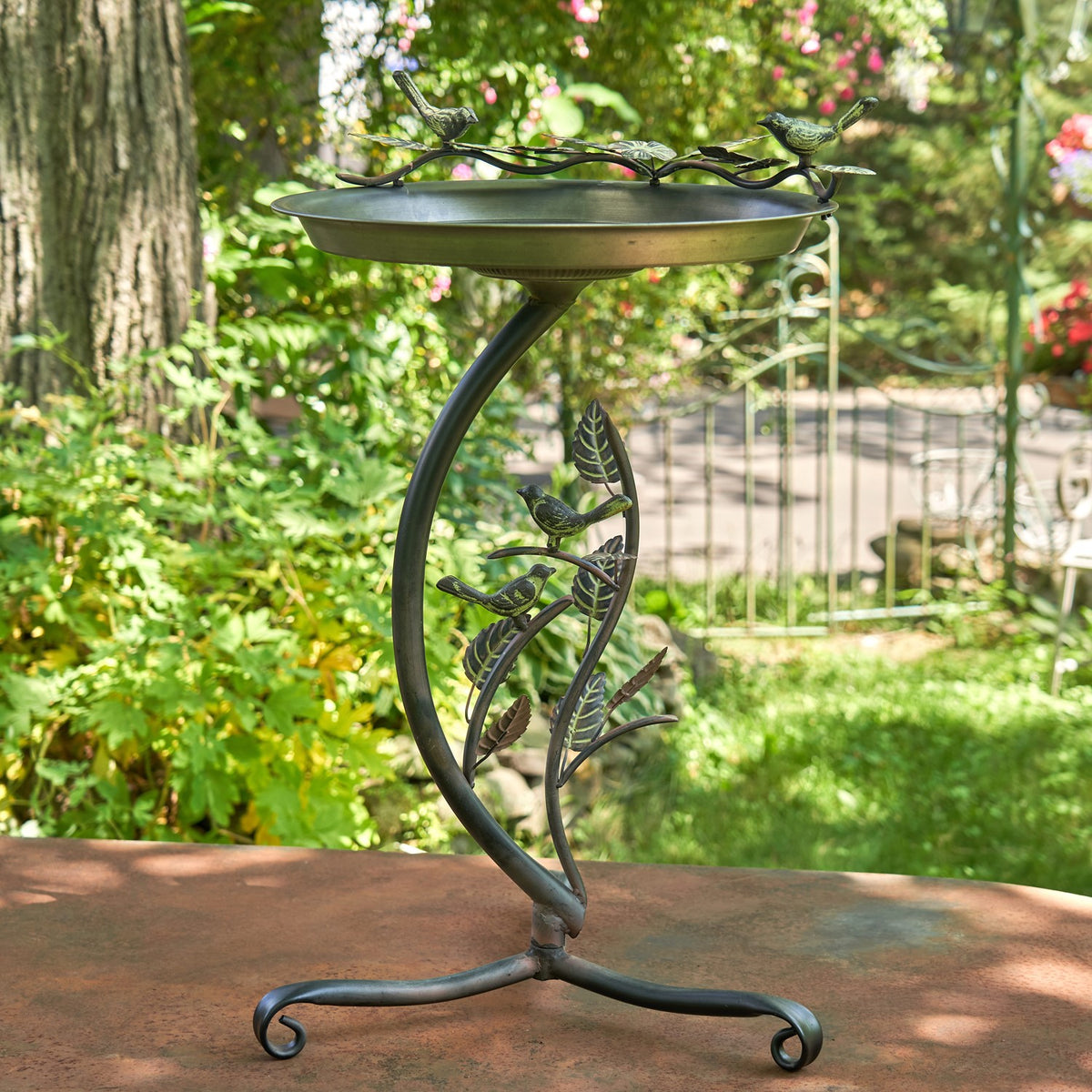 Shallow metal birdbath on a curved branch stand with bird and leaf ornaments, displayed on a garden table.
