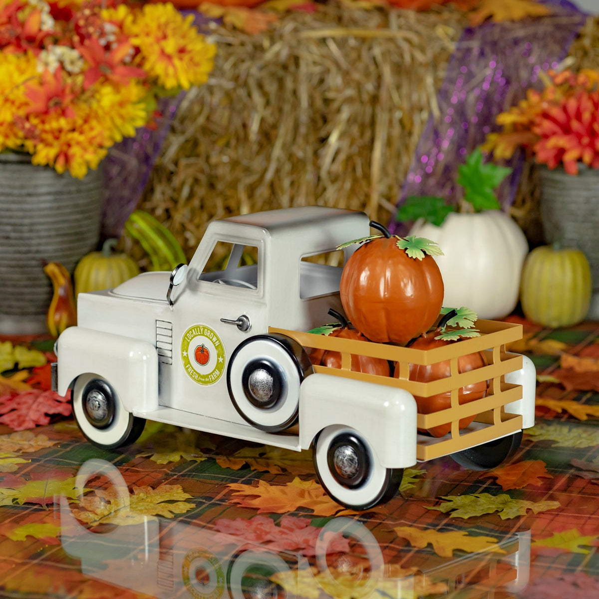 Antique-white miniature pickup truck carrying pumpkins, displayed among autumn leaves and fall decorations.