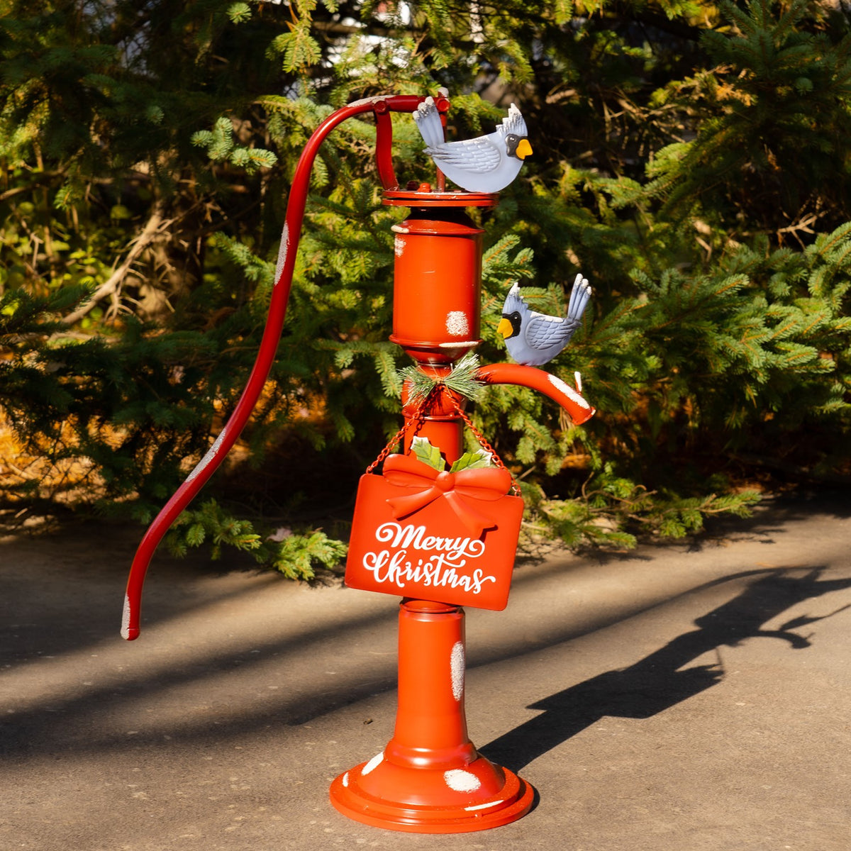 Red vintage-style water pump decoration with two birds and a hanging sign, displayed in front of evergreen branches.