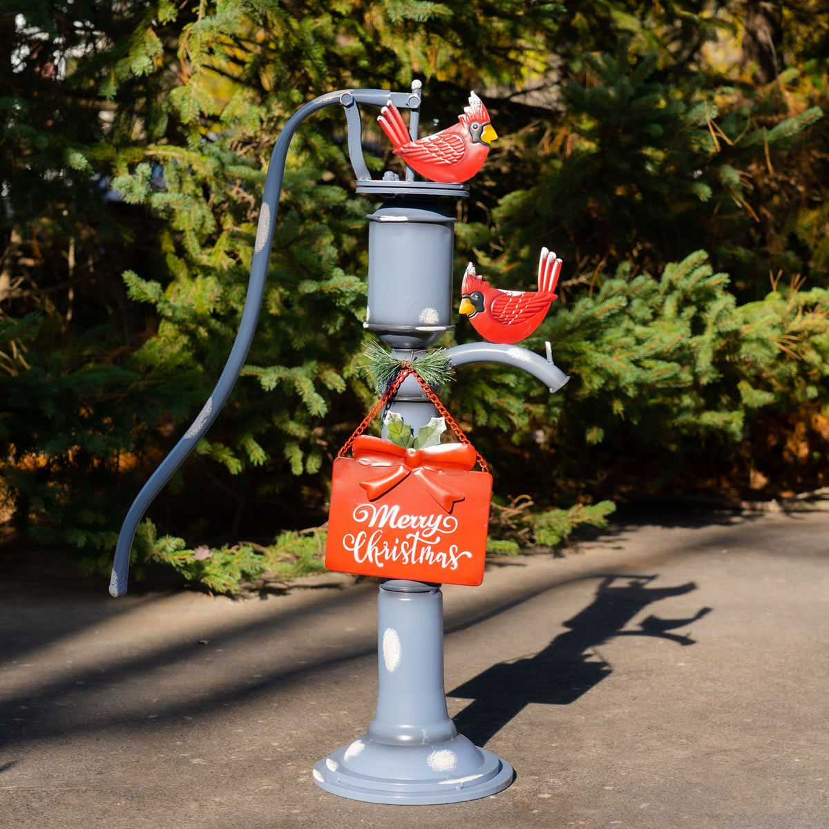 Blue hand pump decoration with two red cardinals and a hanging red Christmas sign, standing outdoors among evergreens.