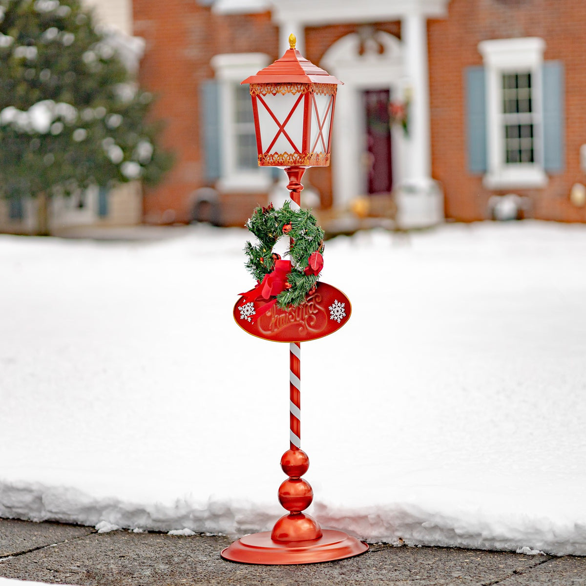 Red standing lantern with a round base, striped pole, and Christmas greenery in a snowy yard.