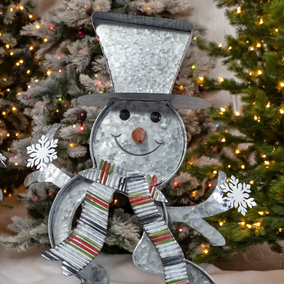 Galvanized metal snowman decoration with a tall hat, striped scarf and snowflake-tipped arms in front of a lit tree.