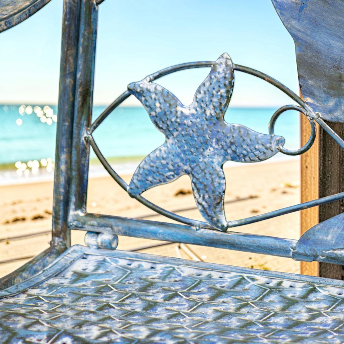 Close-up of a textured metal starfish decoration above the bench’s patterned seat, with beach and sea behind it.