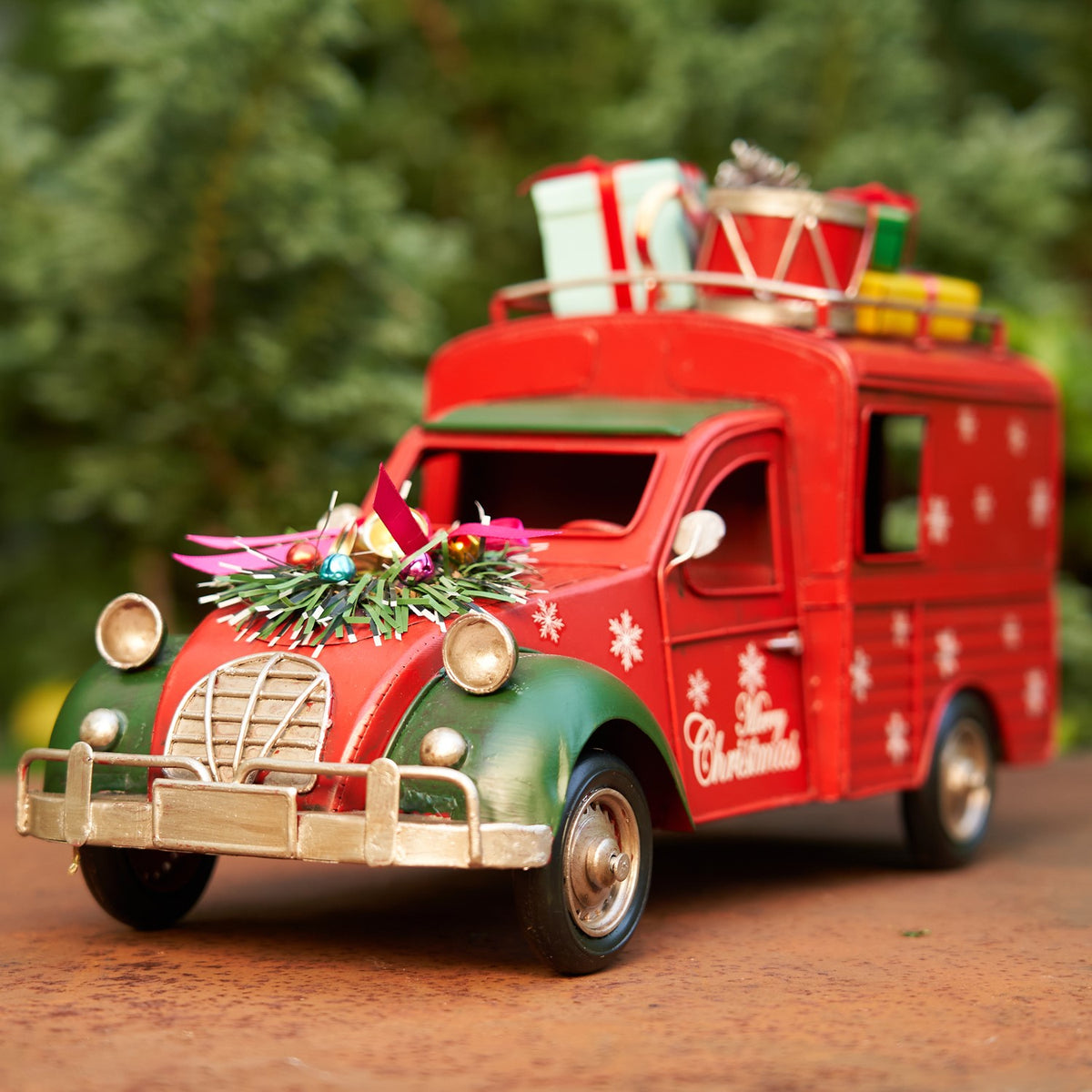 Red vintage-style Christmas truck with gifts on the roof, posed among evergreen branches.
