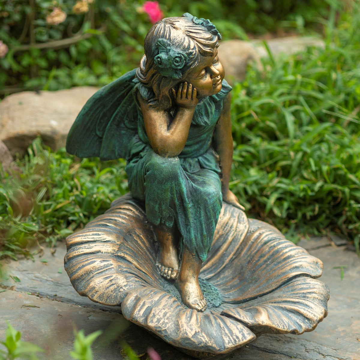 A seated fairy statue rests on a large flower-shaped base amid garden greenery.
