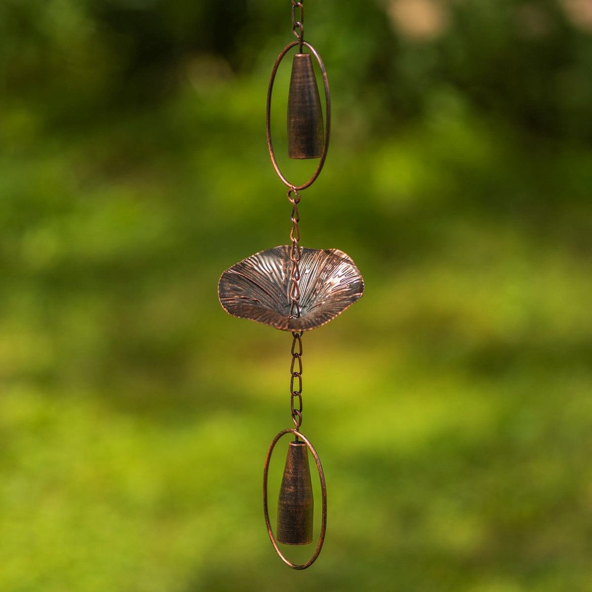Antique bronze rain chain with mushroom-shaped cups and chimes hanging against a leafy green background.