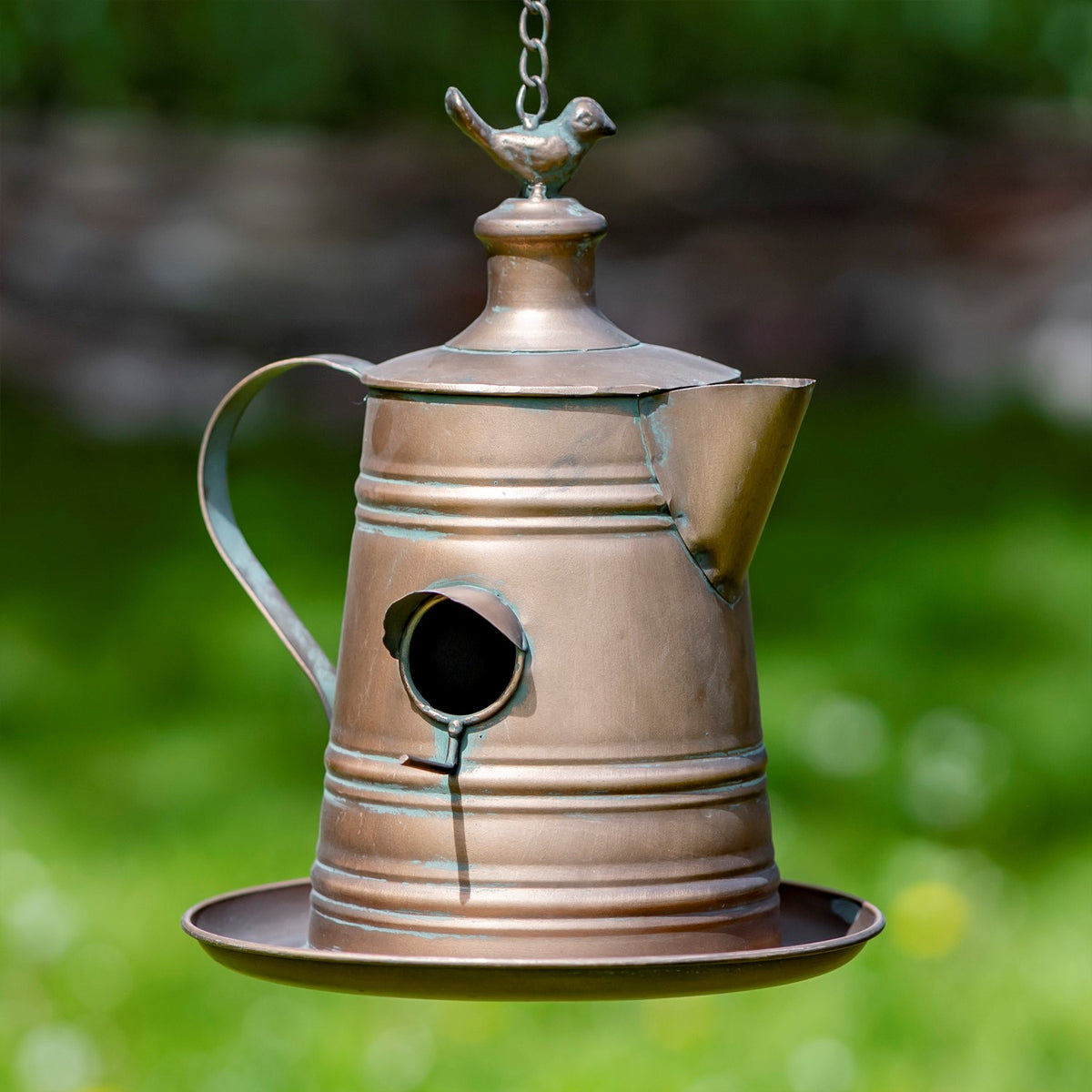 Copper-toned teapot birdhouse hanging from a chain, with a bird finial, round entrance and saucer base.