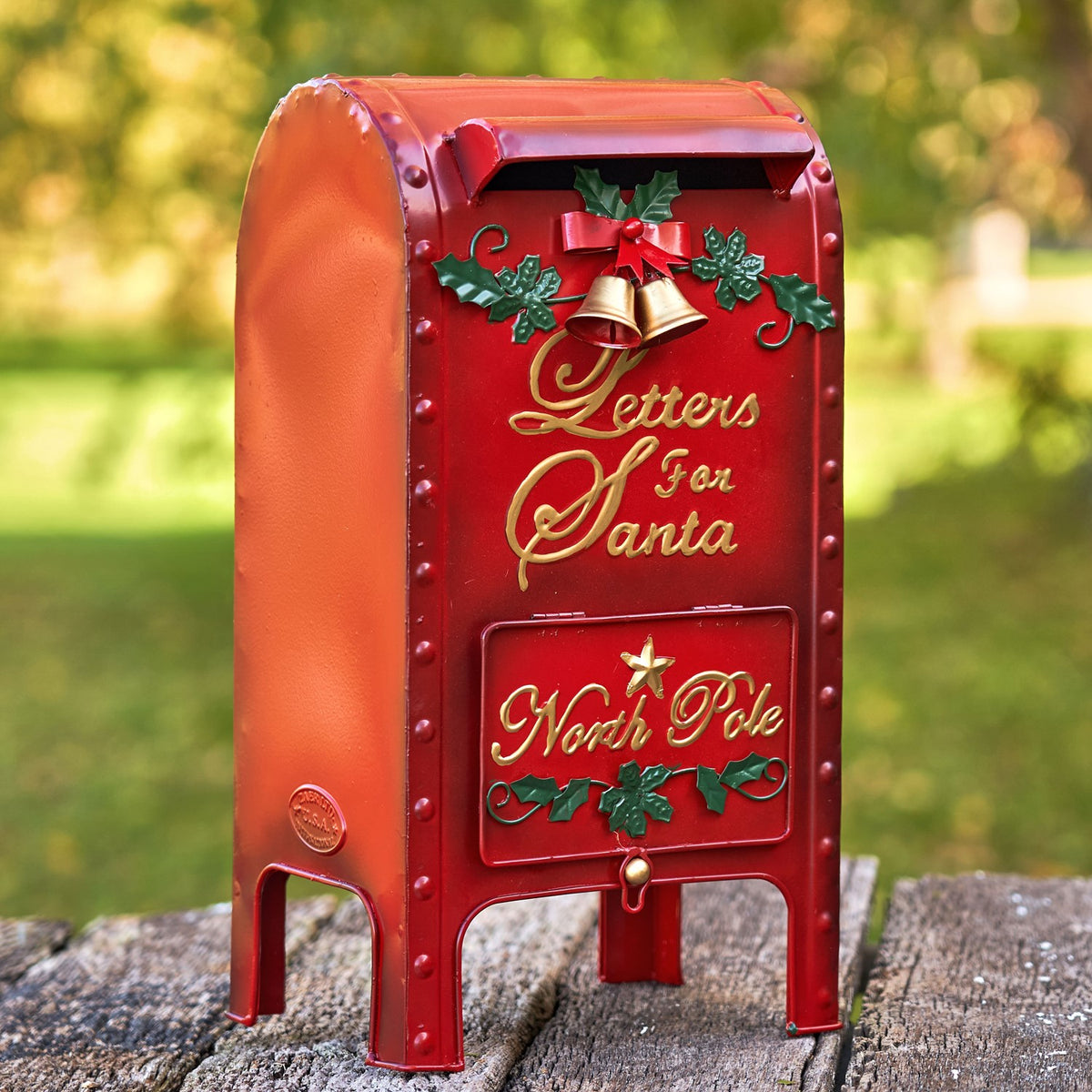 Red holiday mailbox with bells and holly displayed outdoors on a wooden surface.