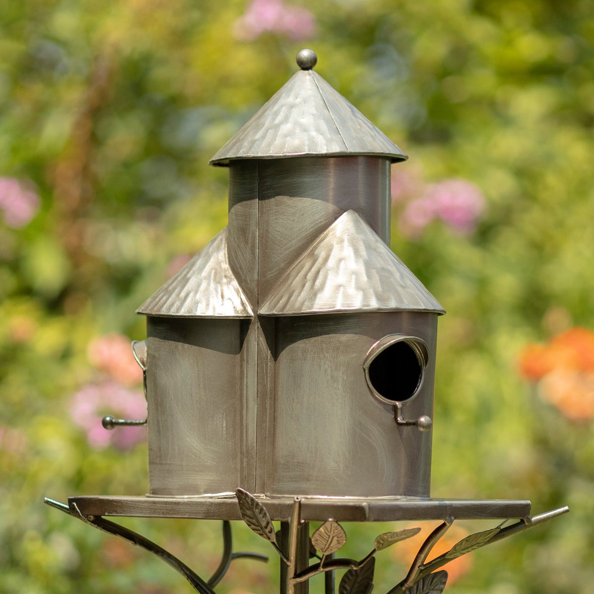 Silver triple birdhouse stake with conical roofs and circular entry holes, shown among garden flowers.
