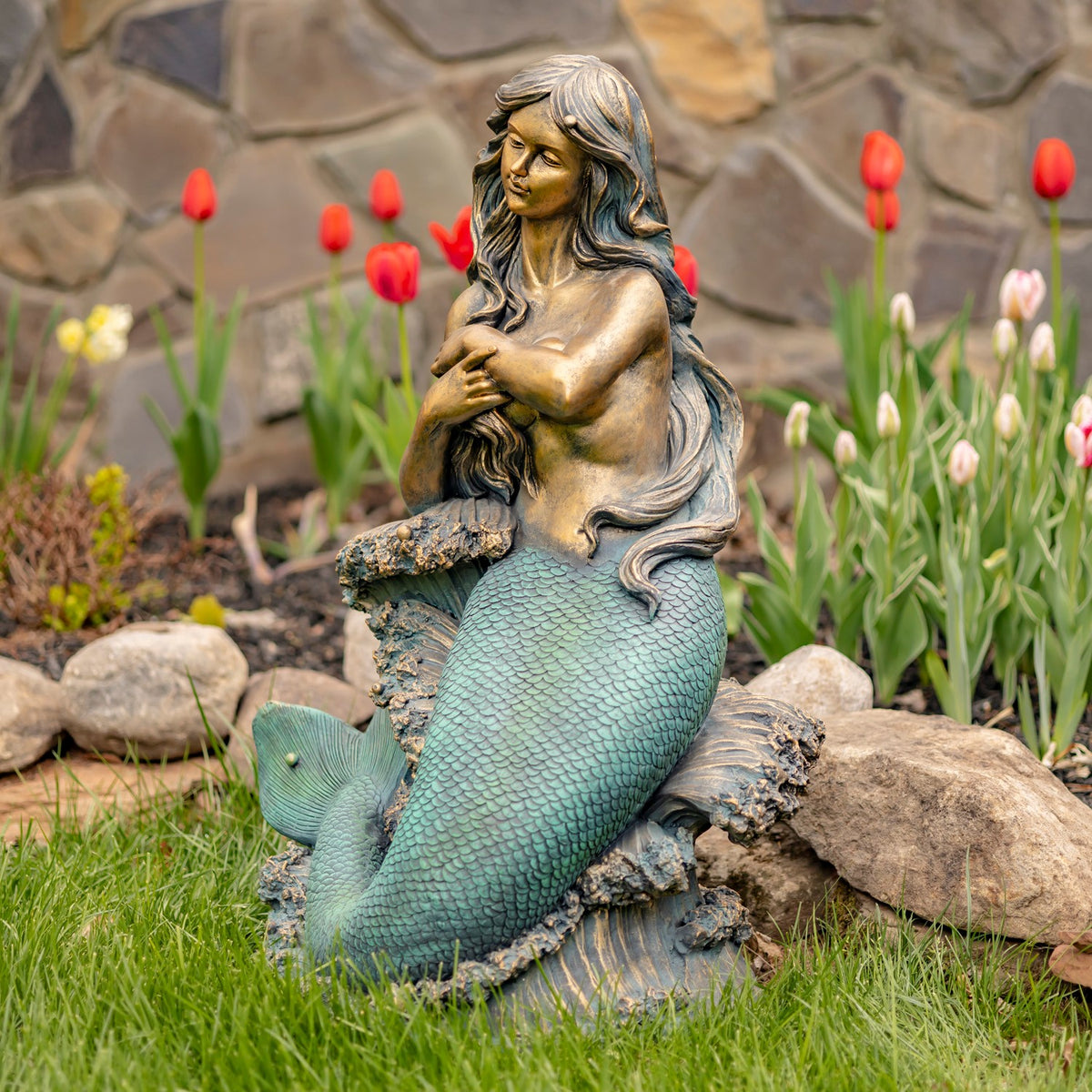 A mermaid statue sits on a textured rock among grass and flowers, facing slightly to the side.
