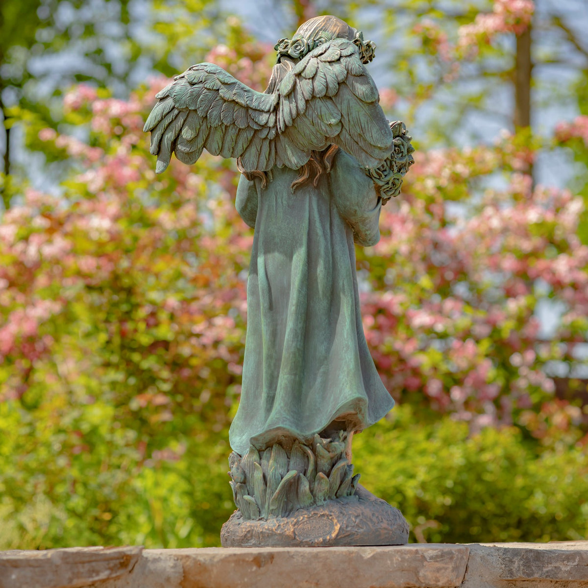 Winged angel statue holding flowers, shown in profile against a garden backdrop.