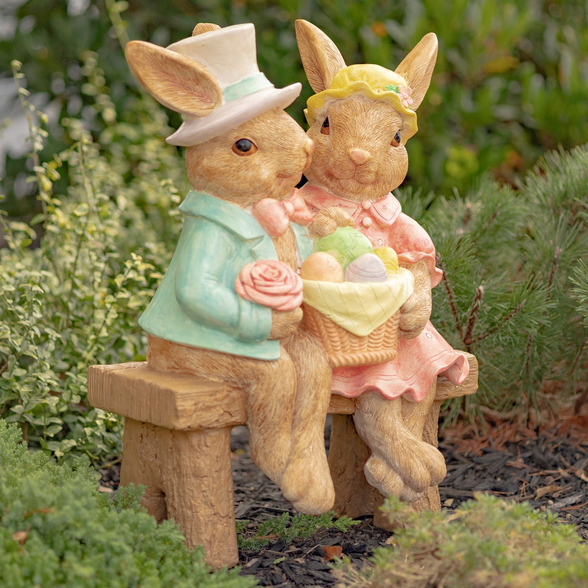 Two dressed rabbits sit together on a rustic bench, holding a basket of colorful eggs among garden plants.