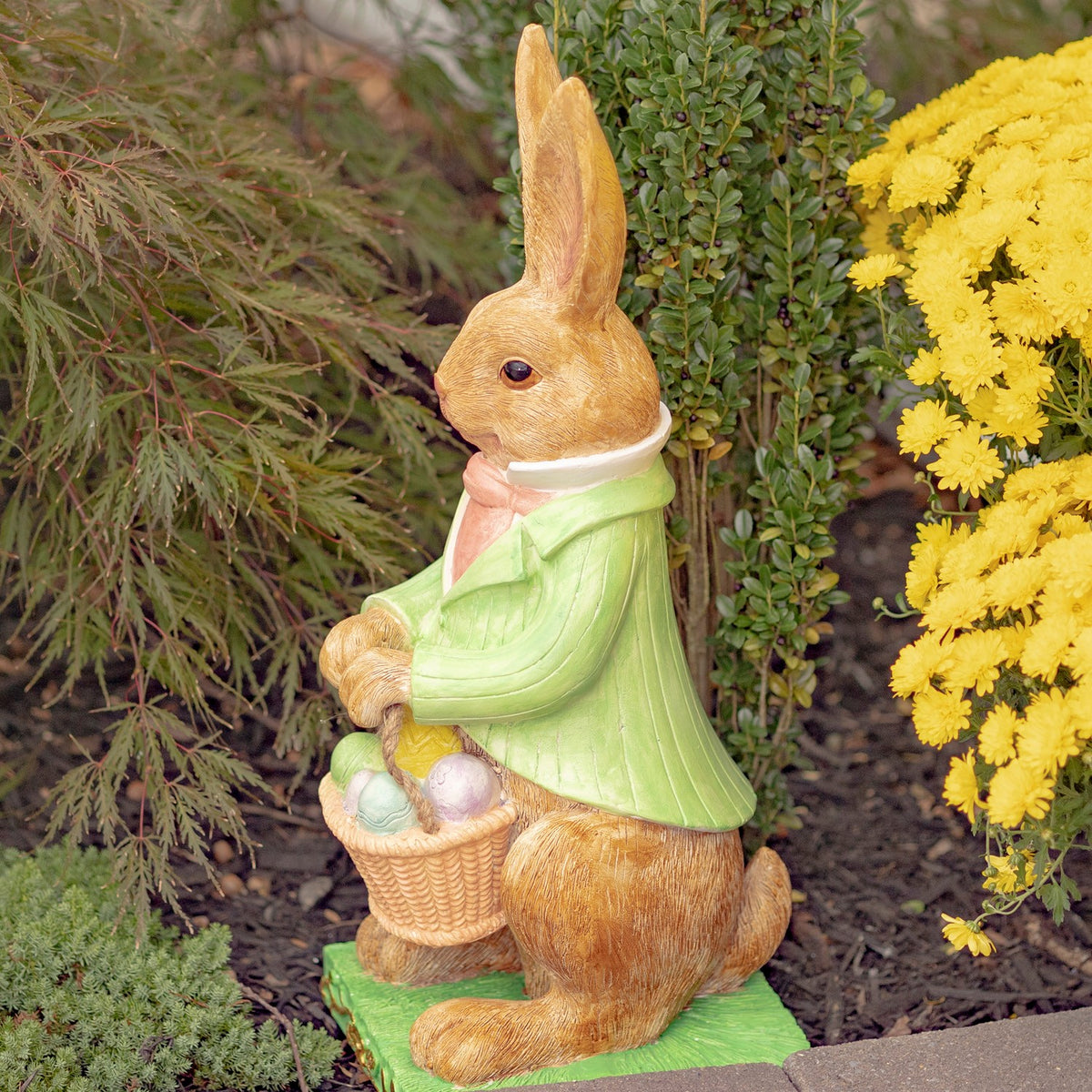 Rabbit statue in a green coat holding an egg basket, displayed among garden greenery and yellow flowers.