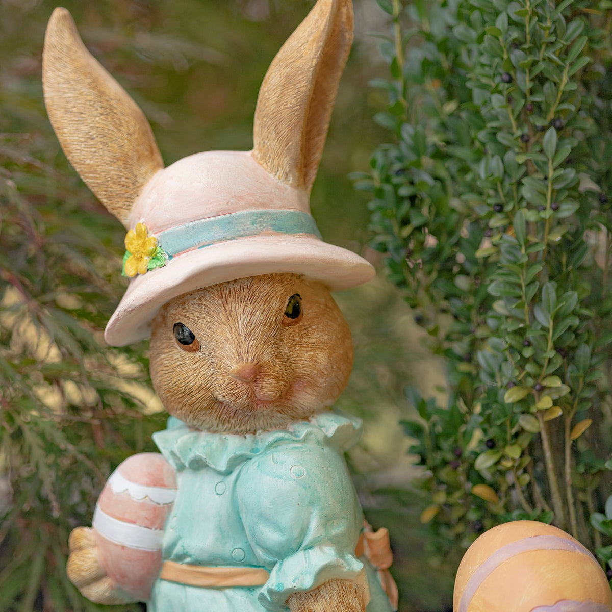 Close view of a rabbit statue wearing a pink sun hat and aqua outfit, with a painted egg beside it.