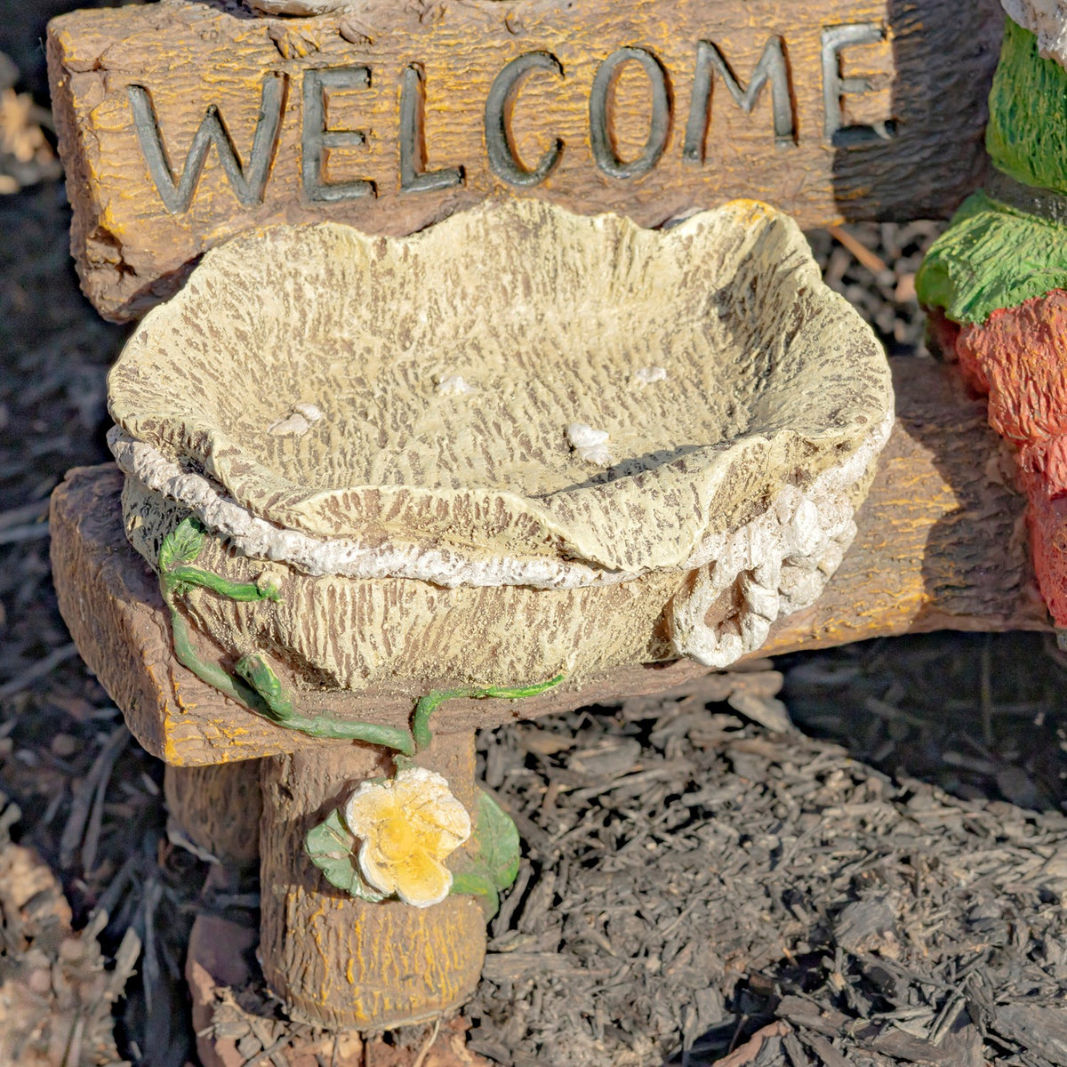 Close view of a textured stone-look birdbath beneath a wooden sign, with part of a colorful gnome at the edge.