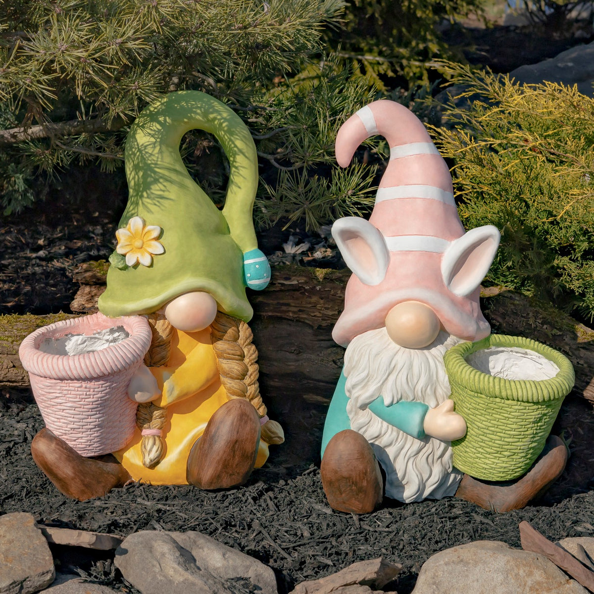 Two seated garden gnomes with colorful hats and basket planters among shrubs and mulch.