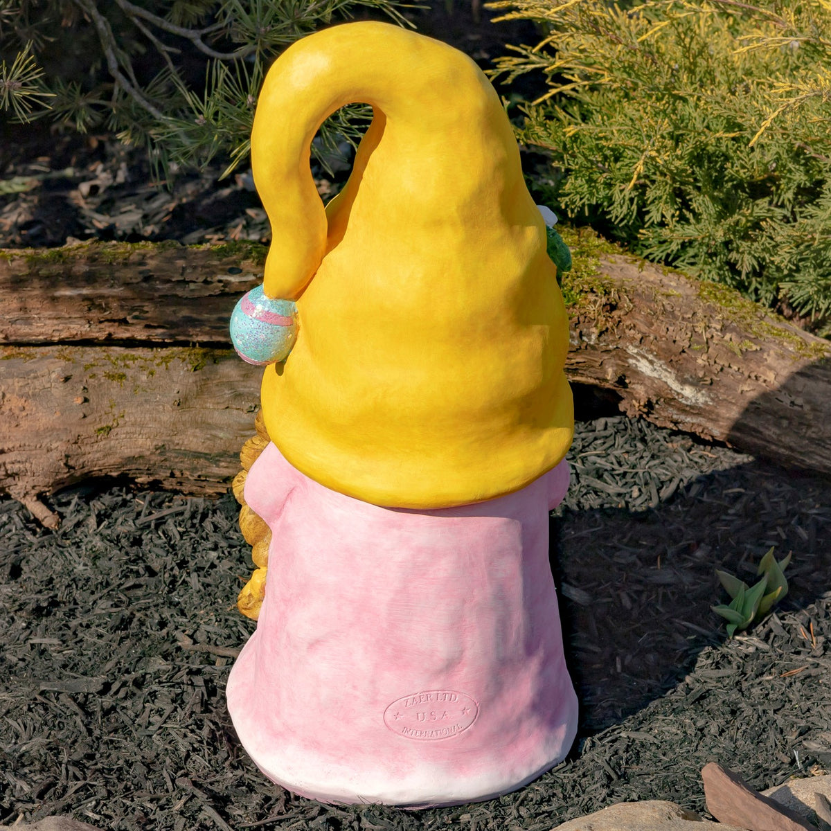 Rear view of the pink gnome with a yellow hat standing in an outdoor garden