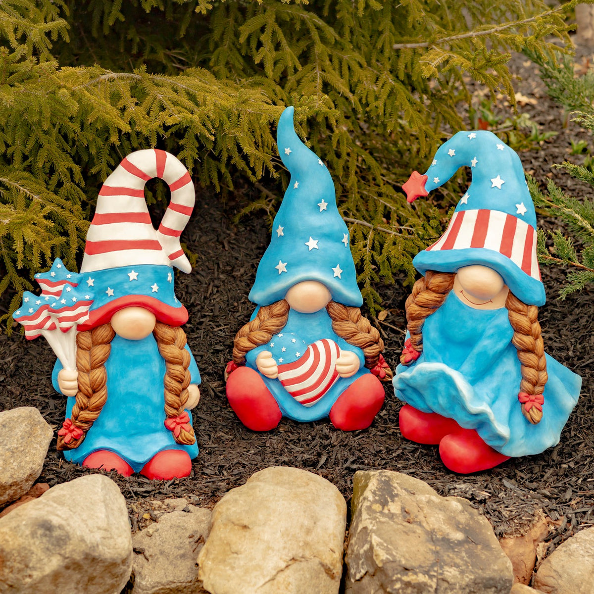Three patriotic lady gnomes sit among garden stones beneath evergreen branches.