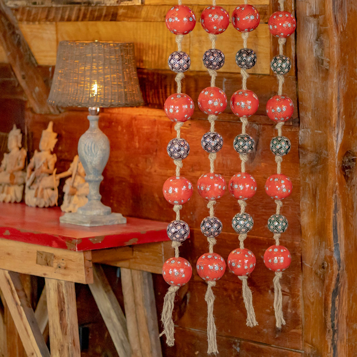 Several strands of red and dark patterned porcelain balls hang against a rustic wood wall beside a lamp.