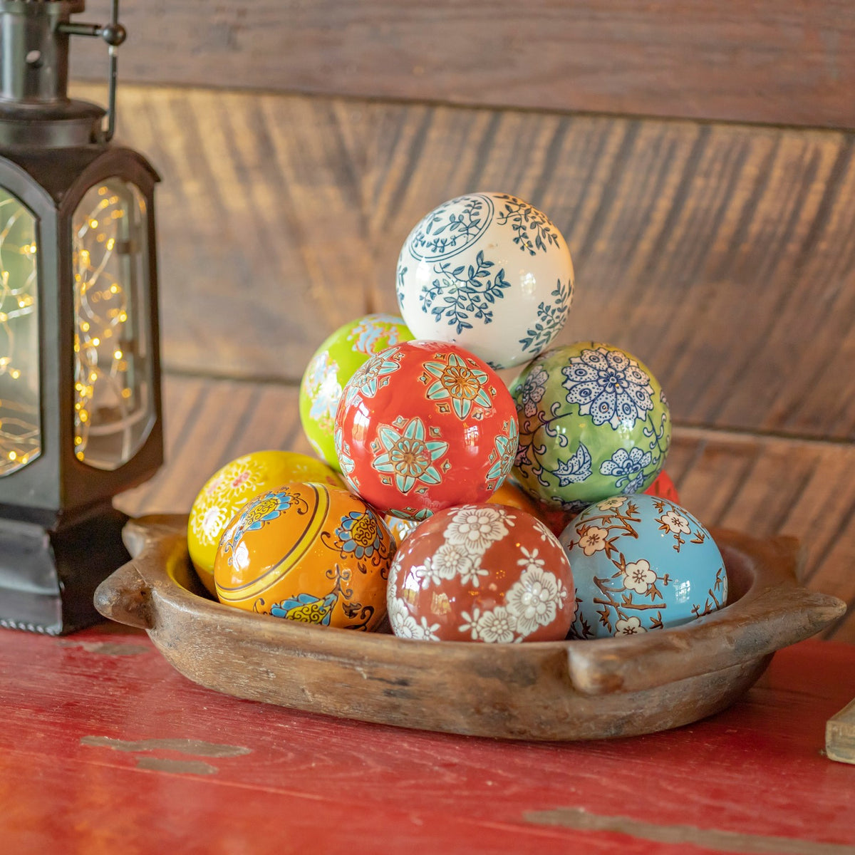 Assorted patterned ceramic balls piled in a wooden bowl beside a lantern.