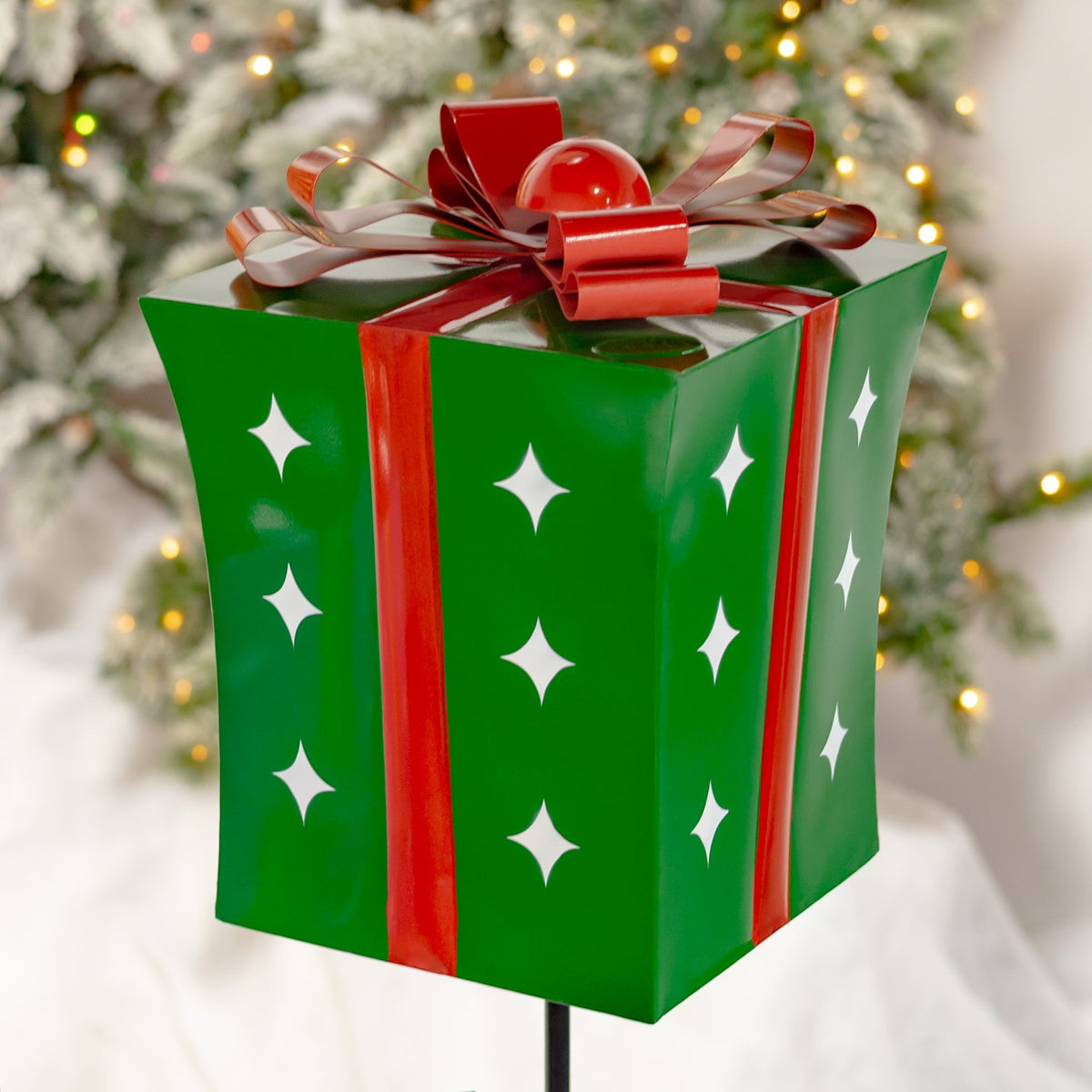 Green gift-box garden stake with a red ribbon and white star cutouts, pictured against a lit Christmas tree.