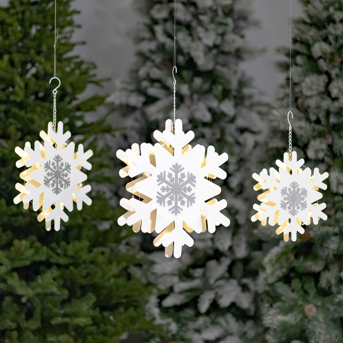 Three white and silver snowflake decorations hanging at different heights before evergreens