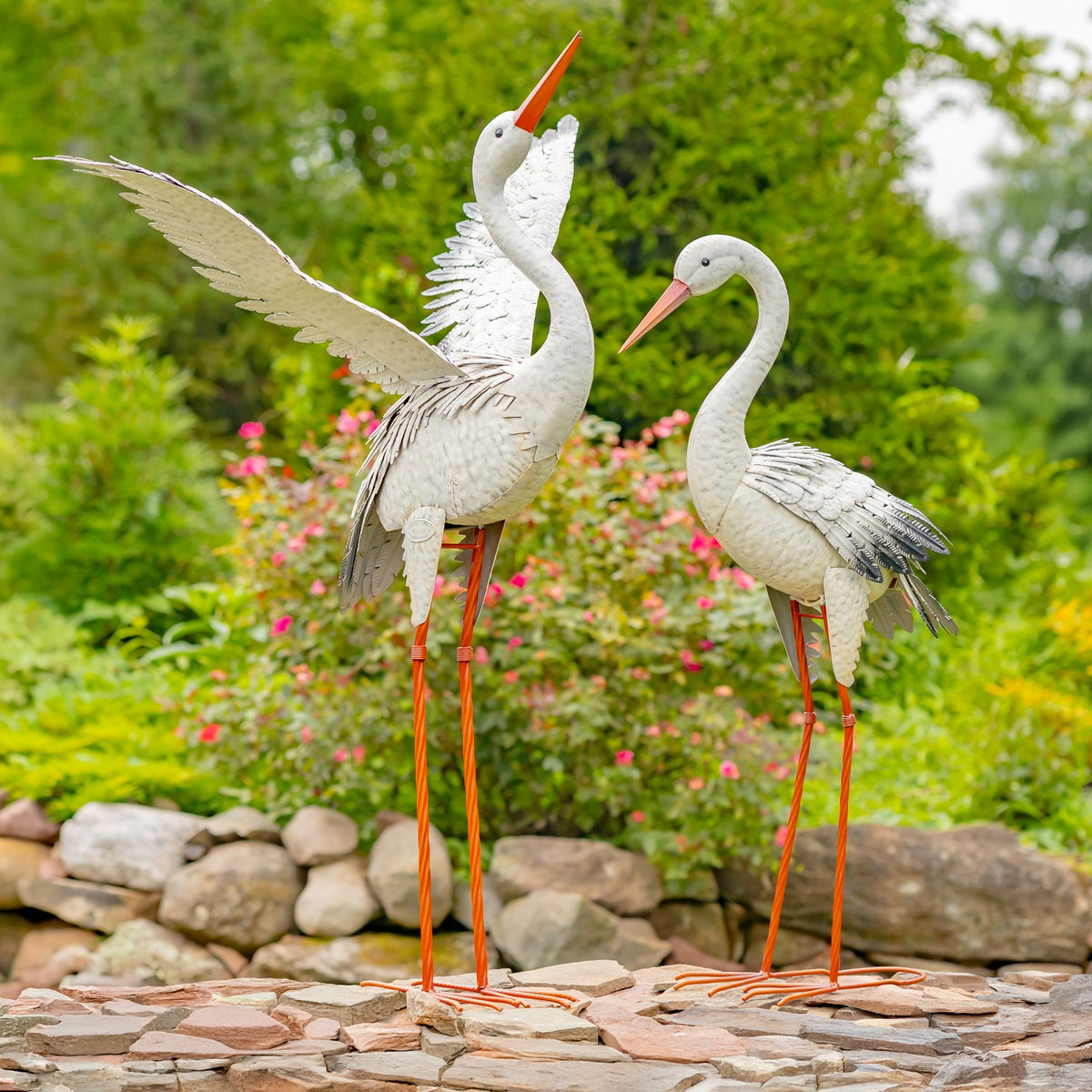 Two white metal heron figures stand on a stone patio, one with a wing extended, amid flowering greenery.