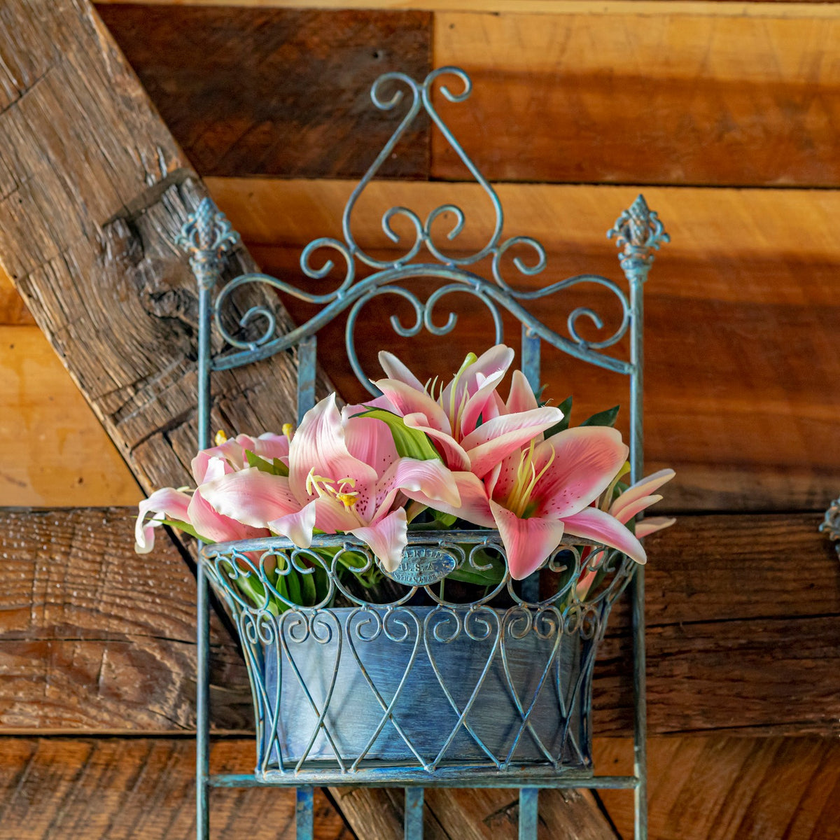 Blue scrollwork wall planter holding pink lilies against a rustic wooden wall.