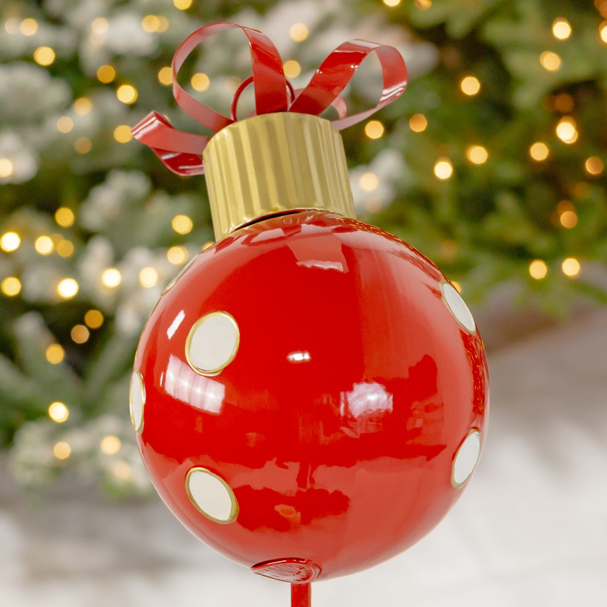 Glossy red Christmas ball with white dots, a gold-toned cap and curled red ribbon against a lit tree.