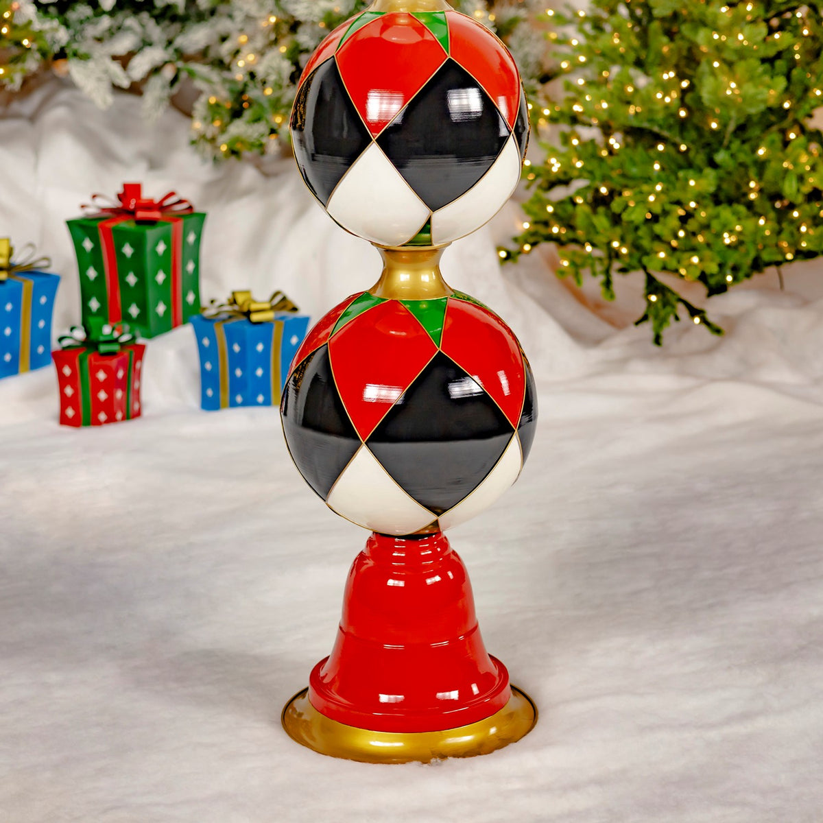A glossy red, black, white, and green stacked ornament tower stands on a gold base beside a lit Christmas tree and presents.