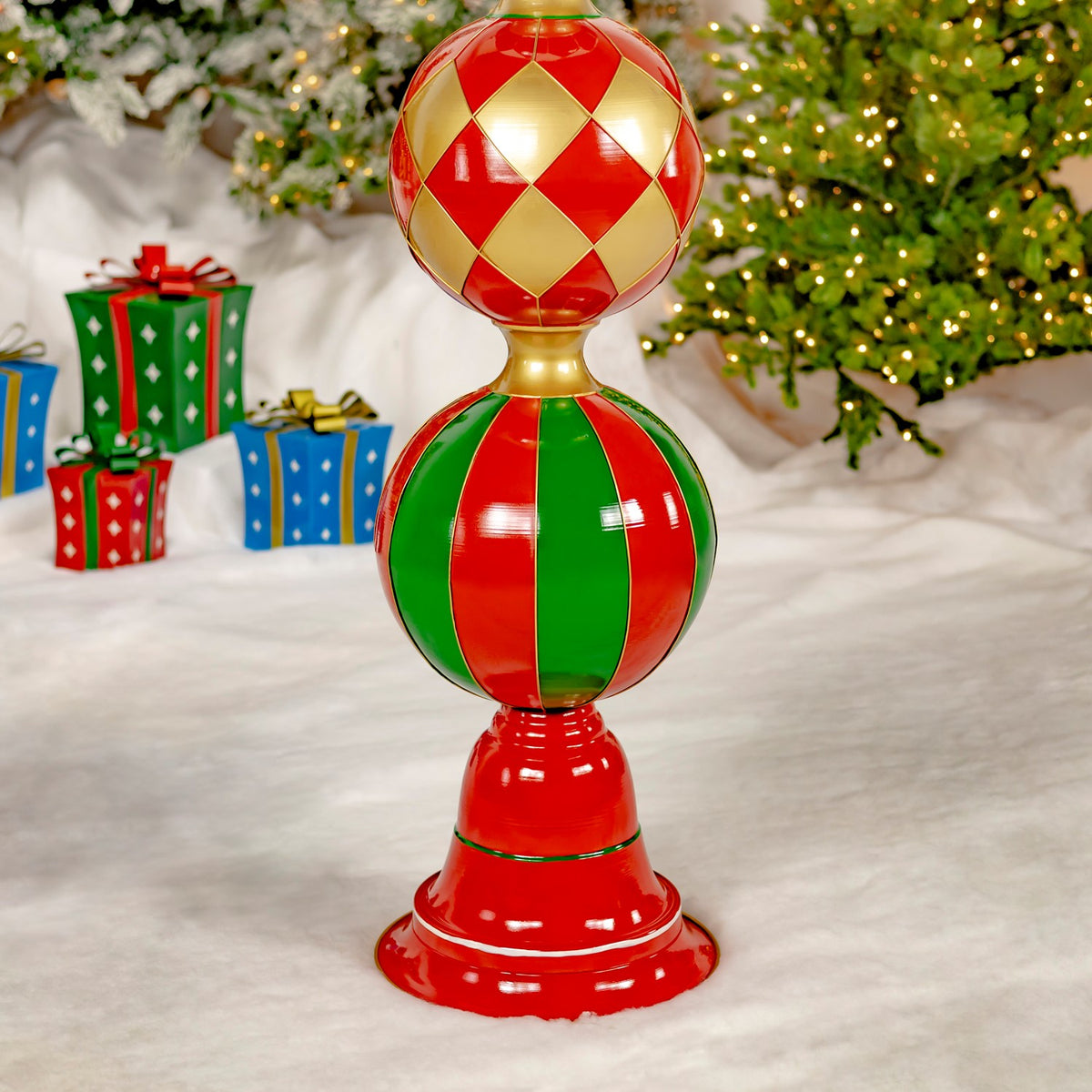 A red, green, and gold ornament tower stands on a snowy floor beneath a lit Christmas tree, with gift boxes nearby.