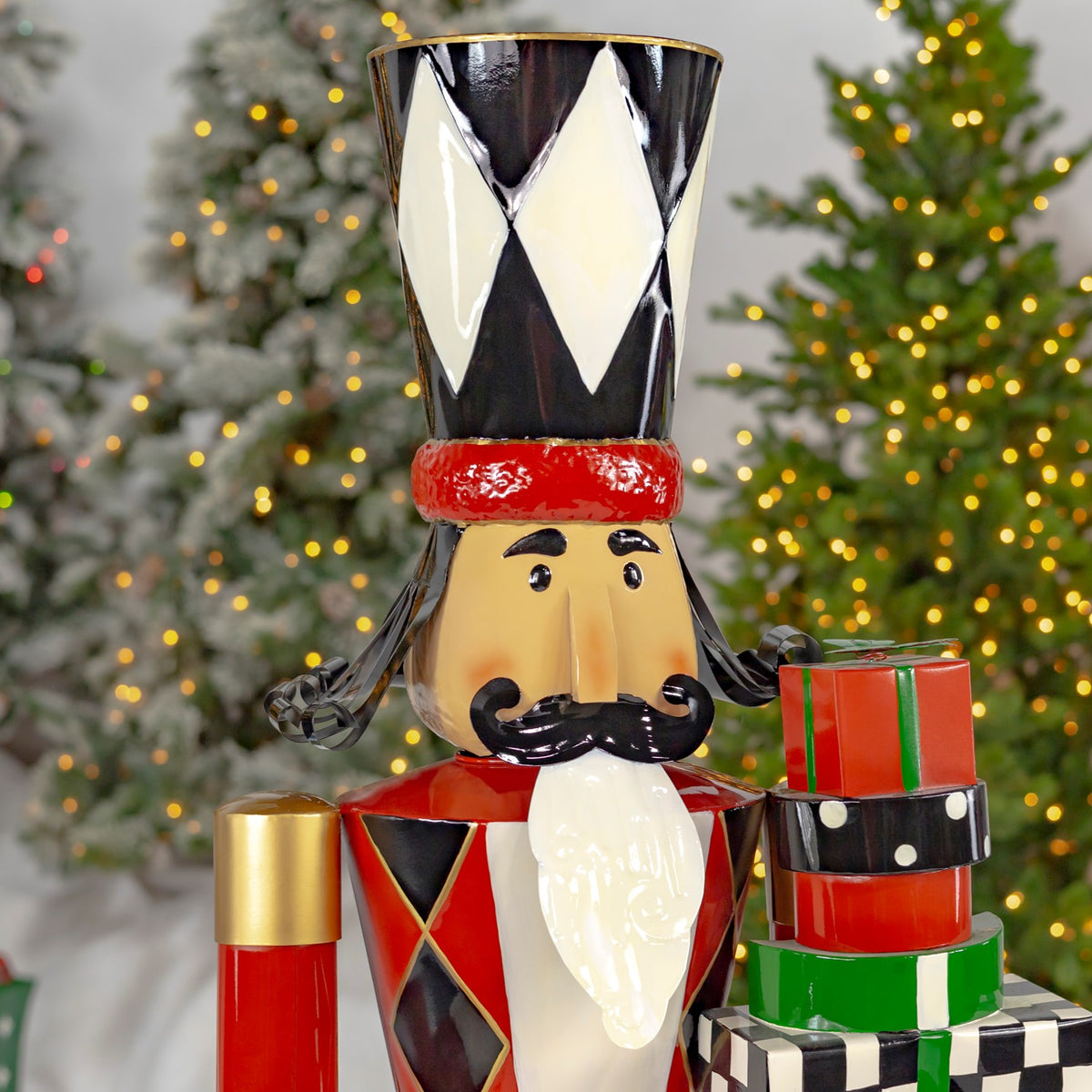 A painted nutcracker holds stacked red and green gifts in front of lit Christmas trees.