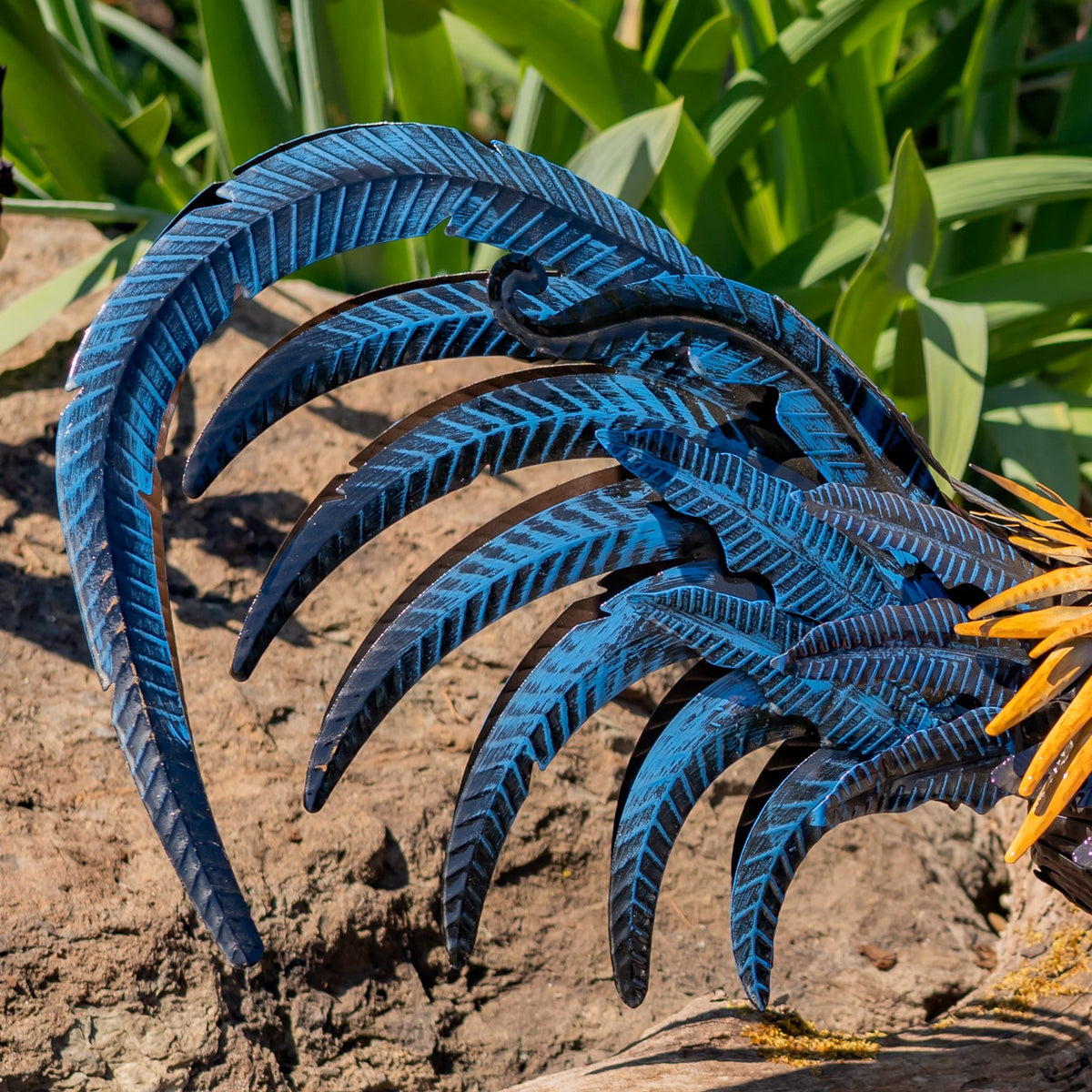 Close-up of the rooster’s layered blue metal tail feathers against a garden backdrop.